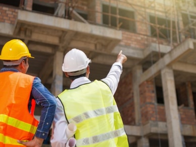 Two men wearing hi-vis vests and hard hats looking up at a construction site, the one on the right is pointing up at the site